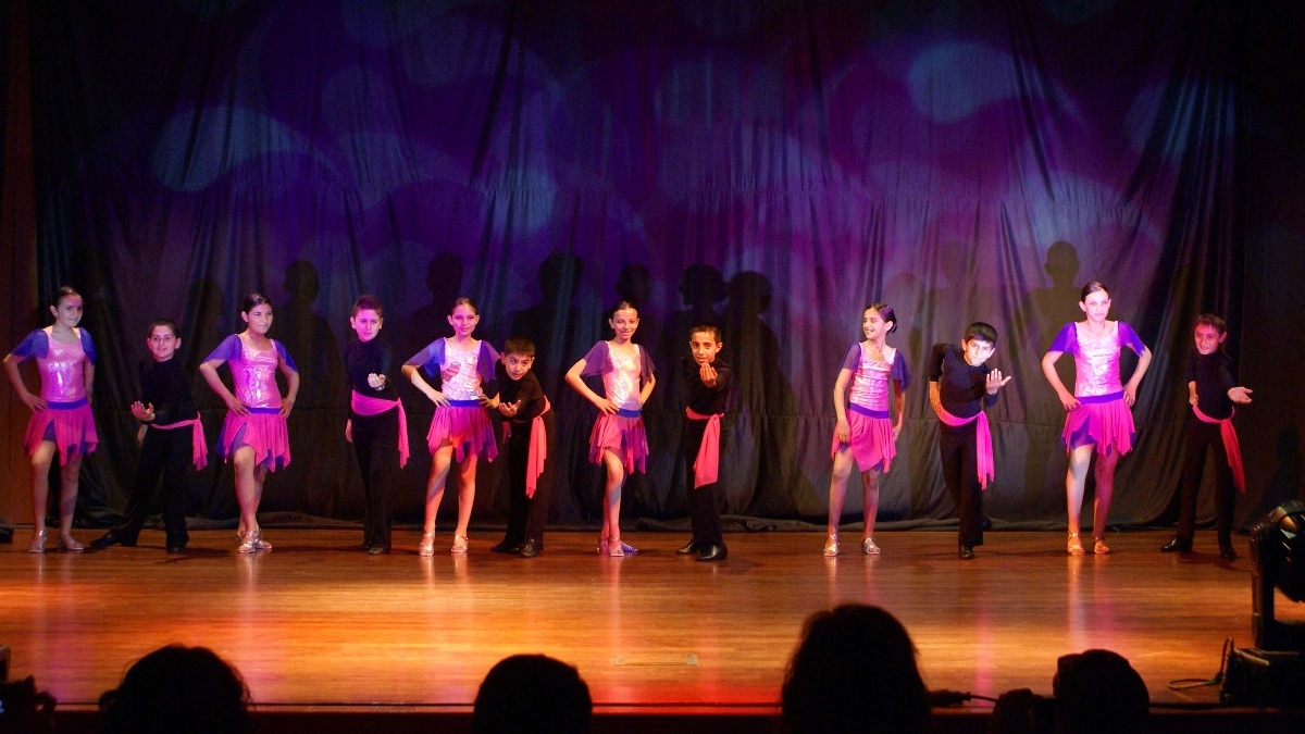 Six couples of children, each couple paired as a girl and a boy, on a stage at an indoors dance recital.